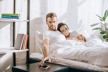 Wall Mural - handsome young man relaxing in bed with girlfriend and reaching for smartphone