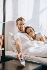 Wall Mural - happy young man relaxing in bed with girlfriend and reaching for smartphone