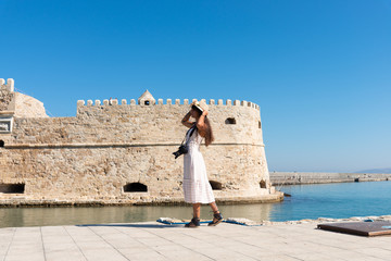 Happy tourist girl on holiday trip to Heraklion, Crete, Greece. Young woman traveller enjoyes sunshine sitting on the dock of Heraklion Venetian port marina. Venetian fort at background.