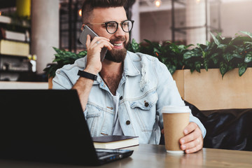 Wall Mural - Young bearded cheerful man sits at table in front of laptops, talking on mobile phone while making notes in notebook.