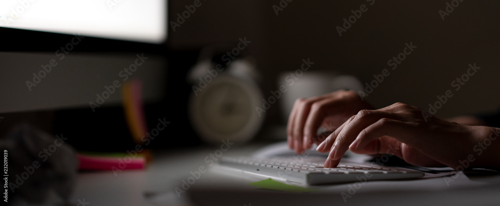 Workaholic hand typing keyboard in dark late night with computer screen light, clock, coffee cup and stuff on desk table with noir working in workplace or hacker cyber crime concept. Work hard banner. - obrazy, fototapety, plakaty 