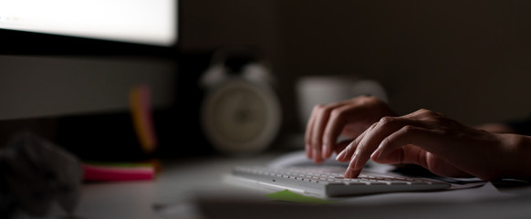 Workaholic hand typing keyboard in dark late night with computer screen light, clock, coffee cup and stuff on desk table with noir working in workplace or hacker cyber crime concept. Work hard banner.