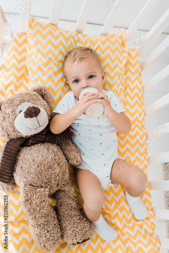 Adorable Toddler In Crib With Fluffy Teddy Bear Toy Drinking Out