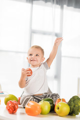 Wall Mural - cute toddler raising hand and sitting on table surrounded by fruit and vegetables