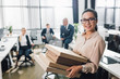 © LIGHTFIELD STUDIOS - young businesswoman holding pizza boxes and smiling at camera while colleagues sitting behind in office