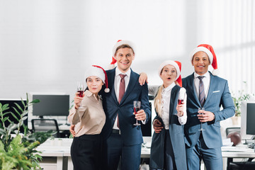 Wall Mural - happy young multiethnic coworkers holding glasses of wine and smiling at camera while celebrating new year in office