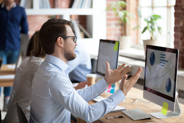 Mad male employee look at screen nervous about computer operation problem, angry man sit in shared office gesturing unhappy about pc malfunction, having slow internet connection or data loss
