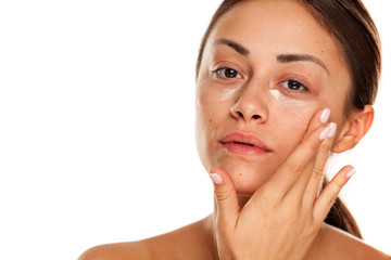 beautiful young woman applying moisturizer on her face on white background