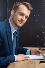 Wall Mural - happy young businessman looking at camera and writing in textbook at table with smartphone in modern office