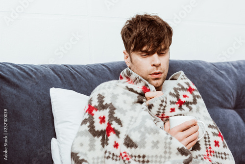 Fotografía young sick man wrapped in blanket drinking tea in bed at home