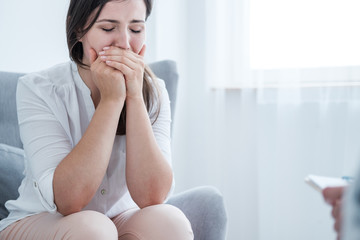 A stressed crying young woman covering her mouth with hands while sitting in a bright room. Empty space in the background.