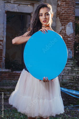 Fényképezés A beautiful girl with long hair in a long black dress with white wings in the image of an angel sits on the stair railing near the walls of an old mansion