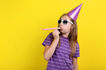 Young girl in birthday hat with sunglasses and whistle on yellow background