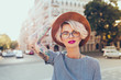 Portrait of pretty blonde girl with short hair posing to the camera on the steet in city. She wears gray checkered  dress, glasses,  tattoo.  She holds hat on head and looks to the camera.