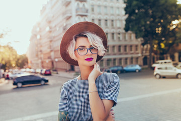 Portrait of pretty blonde girl with short hair posing to the camera on the steet in city. She wears gray checkered  dress, glasses, hat and has purple lips.