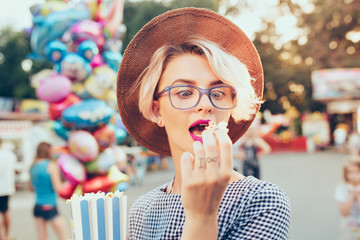 Closeup portrait of  blonde girl with short haircut  outdoor  on baloons background. She wears checkered dress, hat, glasses, purple lips. She is going to eat popcorn.