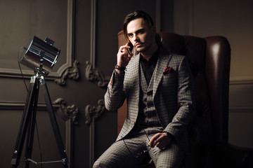 Young handsome hipster sitting in his loft design studio, using phone and communicating with his clients with the help of modern technologies. Fashionable stylish man wearing trendy suit. 