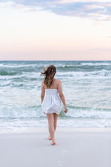 Wall Mural - Young woman in white dress on beach pink sunset in Florida panhandle with wind, ocean waves, walking legs back to water