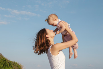 Mother throwing up an adorable baby girl against the clear blue sky, spending together time outdoor, healthy family lifestyle
