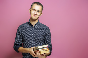 Young man holding a stack of books on a pink background.