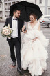 © sonyachny - Gorgeous bride and stylish groom walking under umbrella in rainy street and smiling. Sensual wedding couple embracing. Romantic moments of newlyweds. Modern  wedding photo