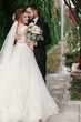 © sonyachny - Gorgeous bride and stylish groom gently hugging on background of green trees. Sensual wedding couple embracing. Romantic moments of newlyweds. Modern wedding photo