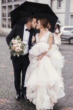 Gorgeous Bride And Stylish Groom Walking Under Umbrella In Rainy Street And Kissing. Sensual Wedding Couple Embracing. Romantic Moments Of Newlyweds. Modern  Wedding Photo