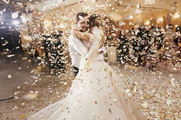 gorgeous bride and stylish groom dancing under golden confetti at wedding reception. happy wedding c
