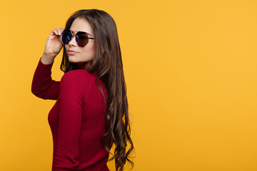 Wall Mural - Pretty young lady in red dress and sun glasees is posing with hand on face on orange background.