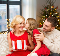 Poster - holidays, family and celebration concept - happy mother, father and daughter with gift at home over christmas tree lights background