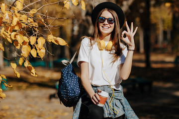 Image of a bright smiling hipster girl with brown hair wearing a hat, sunglasses and backpack holding cup of tea or coffee in the park