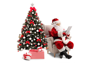 Santa Claus sitting in an armchair next to a Christmas tree and using a tablet