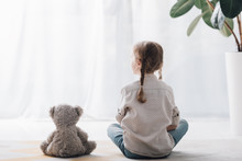 Rear View Of Little Child Sitting On Floor With Teddy Bear And Looking Away
