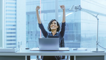 Shot Of The Beautiful Businesswoman Sitting At Her Office Desk, Raising Her Arms In A Celebration Of A Successful Job Promotion.