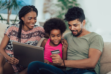 Cute little Afro-American girl and her beautiful young parents using a laptop and doing shopping online while sitting on a sofa at home.