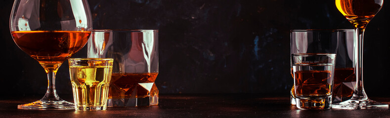 Set of strong alcoholic drinks in glasses and shot glass in assortent: vodka, rum, cognac, tequila, brandy and whiskey. Dark vintage background, banner, selective focus