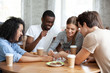 © fizkes - Diverse multiracial friends cheerful girls and guys drinking coffee having fun laughing assembling puzzle jigsaw sitting together at table in home or cafeteria. Teamwork and weekend activities concept