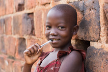 An 11-year Old Ugandan Girl Smiling, Holding A Pen Against Her Mouth And Leaning Against A Brick Wall Looking At The Camera