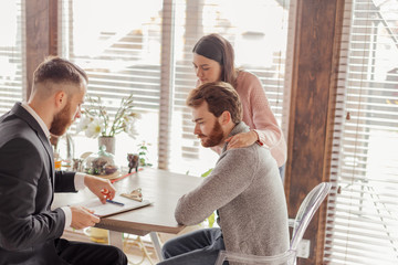 Young caucasian couple consulting with bank financial adviser before buying new house. Two bearded men sitting at table and checking documents while woman stands behind.