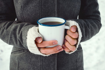 Closeup of woman's hands holding white enamel cup with hot drink outdoors, selective focus