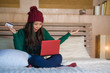 © TheVisualsYouNeed - young beautiful happy Asian Korean woman in winter hat relaxed on bed holding credit card using laptop computer for internet banking and online shopping