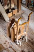 Vintage Hand Barrow Standing Upright On Wooden Floor, In Flour Mill, With Two Large Bags Of Flour In Front, Floor Is White From Flour Dust