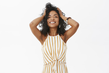 Wall Mural - Girl feeling beautiful and feminine today. Charming happy and delighted stylisht tender african american female playing with curly hair and smiling satisfied at camera posing over gray wall