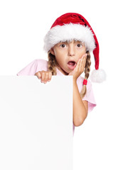 Wall Mural - Portrait of surprised little girl in Santa hat with white blank. Child wearing Santa Claus hat, posing behind white panel. Kid holding empty Christmas billboard, peeping behind blank holidays board. 