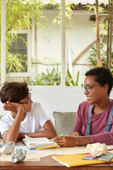 Wall Mural - Professional dark skinned designer explains how make project work to male trainee, has pleasant smile on face, pose together in outdoor cafeteria in exotic resort place, points with pen into diary