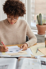 Wall Mural - Vertical shot of hard working student with Afro haircut, checks text documentation, prepares pie chart for describing statistics, writes in documents, wears casual sweater. Woman carries accounting