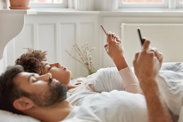 Horizontal shot of serious multiethnic young woman and man focused in modern gadgets, monitor web multimedia content, lie in bed, connected to wireless home internet, share files, message online