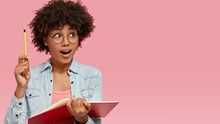 Isolated Shot Of Surprised Clever Student With Intelligent Look, Raises Hand With Pencil, Has Good Idea For Writing In Essay, Holds Red Notebook, Wears Fashionable Clothes, Poses Over Pink Background