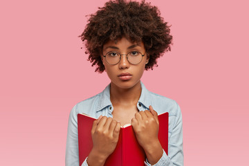 Wall Mural - Photo of serious attractive bookworm holds red textbook, reads sad story, wears round spectacles, isolated over pink studio wall. Horizontal shot of clever schoolgirl enjoys reading, poses indoor