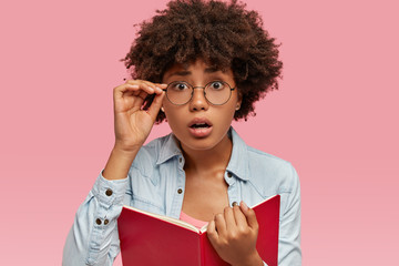 Wall Mural - Image of surprised black woman has Afro haircut, cant believe in bad exam result, wears round glasses, carries textbook, dressed in fashionable clothes, isolated over pink background. Omg concept